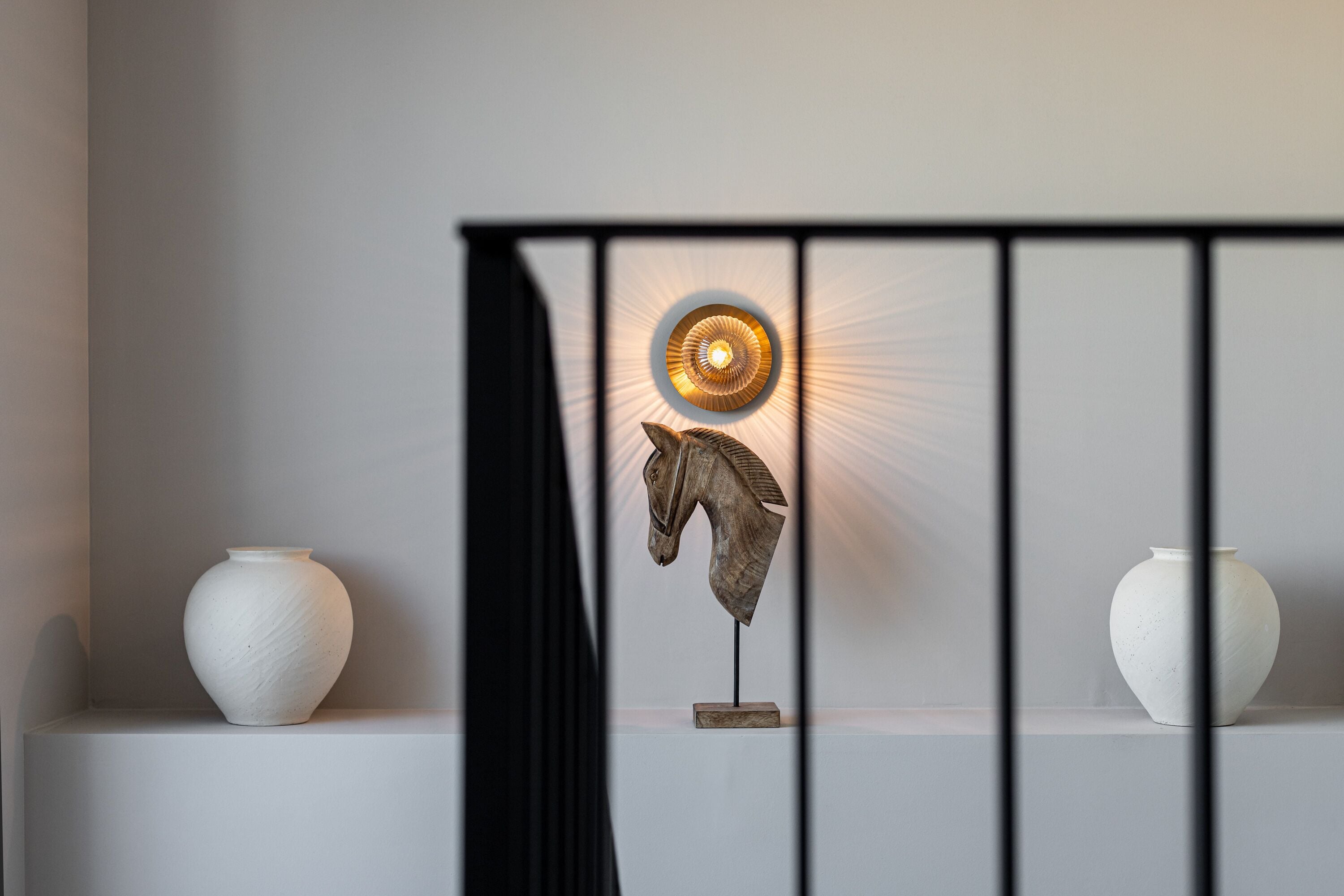 Une pièce avec un vase blanc posé sur une étagère, une sculpture décorative représentant une tête de cheval et un luminaire fixé au mur.