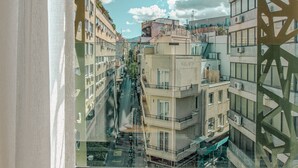 A view through a window with a geometric frame, revealing a narrow urban street lined with multi-story buildings, some with balconies and air conditioning units. - The Residence Aiolou Hotel & Spa (Athens)