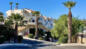 A multi-story building with a white facade and red-tiled roof, surrounded by palm trees and greenery.