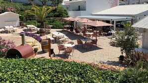 An outdoor patio area with tables and chairs, surrounded by greenery and a white building.