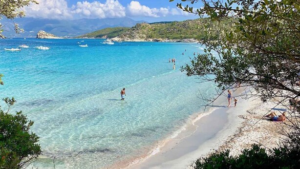Uma praia com águas turquesa cristalinas, uma faixa de areia e barcos ao longe.