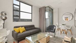 Una sala de estar moderna con un sofá gris, una mesa de centro de vidrio y un comedor con sillas blancas y una mesita.