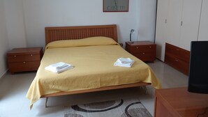 En snyggt bäddad säng med ett gult överkast, två sängbord, ett litet bord, en byrå i trä och ett inramat konstverk på väggen.