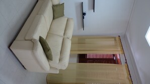 Ett vardagsrum med en beige soffa, en grön kudde, en väggmonterad TV och tunna, genomskinliga gardiner.