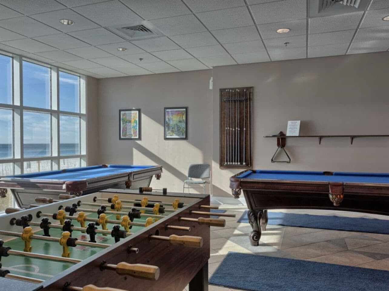 A room with two foosball tables, a pool table, and a window providing a view of the outdoors.