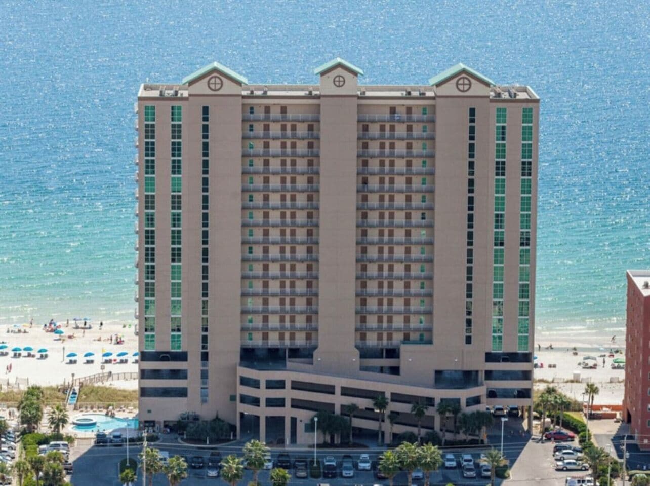 Un hotel de varios pisos con un patio central, situado cerca de una playa con sombrillas y palmeras.
