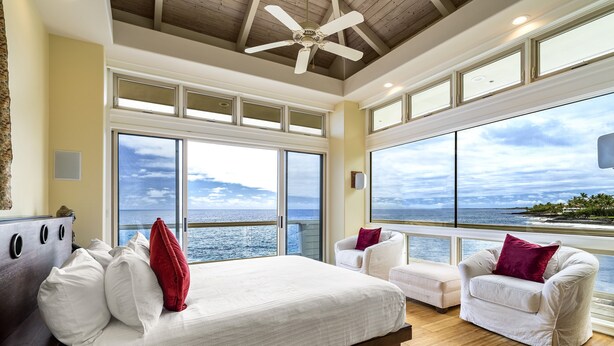 Un dormitorio amplio con una cama grande, dos sillones blancos y vistas al océano a través de amplias ventanas.