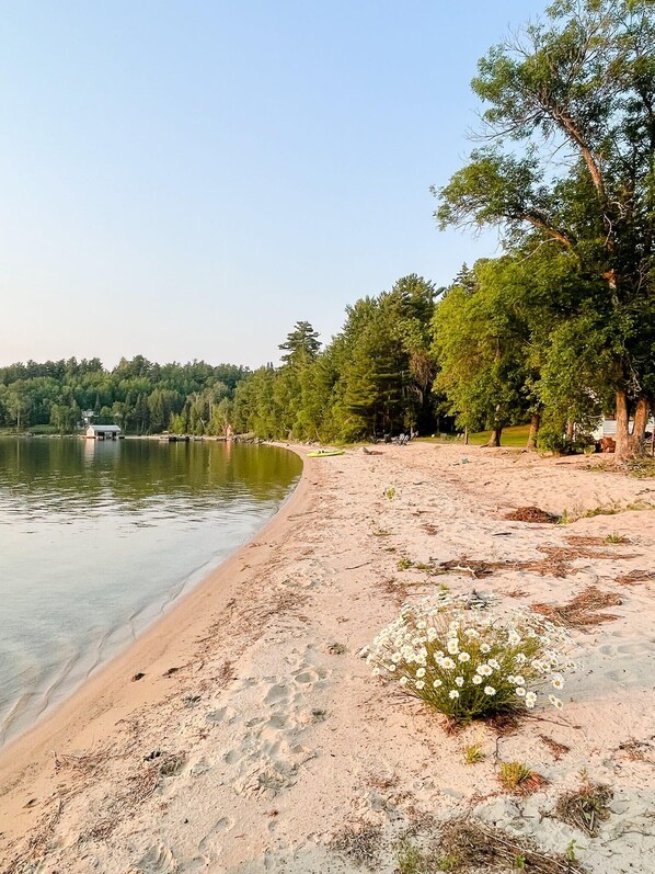 Une plage de sable bordée d’un lac paisible, entourée d’une forêt dense et, au loin, de quelques bâtiments.