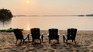 A beach with four chairs facing the water, a small table, and a green kayak. - Bowerbird Beach Camp Cabin #2 (Sioux Narrows-Nestor Falls)