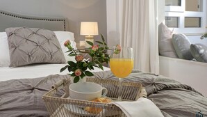 Ein ordentlich gemachtes Bett mit grauem Kopfteil, ein Nachttisch mit Lampe, eine Ablage mit Orangensaftglas, einer Schüssel und ein Korb mit einem Croissant und einer Rose.