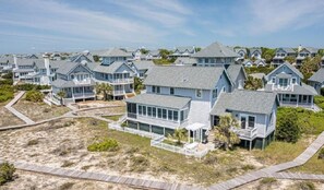 A coastal residential area with houses featuring large windows, some with balconies, and a sandy landscape with wooden walkways.
