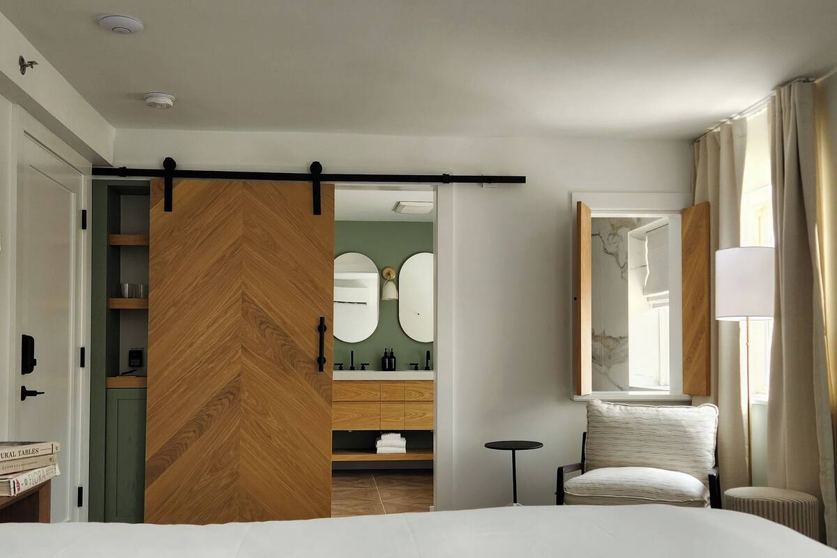 A hotel room with a large bed, a book on the bed, a chair, a small table, a mirror, and a sliding door leading to a bathroom.
