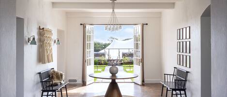 A spacious room with a round table at the center, two chairs on either side, a chandelier, and a view through an open door to an outdoor area with a garden and a tent.