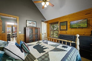 A bedroom with a wooden ceiling fan, a large bed with a quilt, a dresser, and a television on the wall.