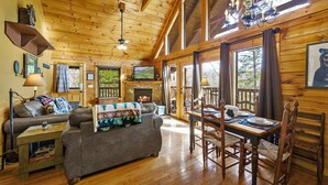 L’intérieur chaleureux d’un chalet avec un plafond en bois, une cheminée, une table à manger avec des chaises et un canapé recouvert d’une couverture.