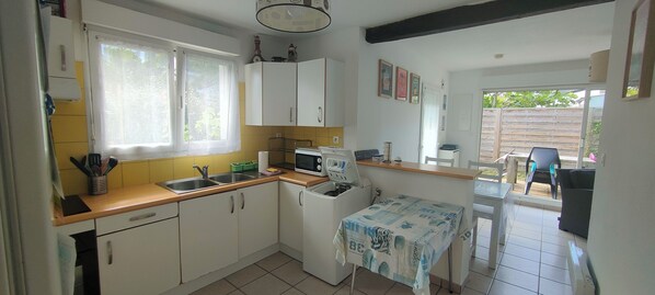 A compact kitchen with white cabinets, a microwave, a small refrigerator, and a sink. There is a window providing natural light, and a table with a tablecloth is visible in the foreground.
