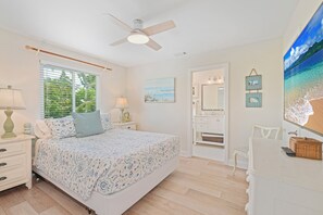 Une chambre à coucher comprenant un lit, des tables de chevet, un ventilateur de plafond, une fenêtre avec des stores, un tableau, un miroir et une commode.