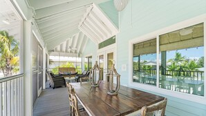 Ein überdachter Außenbereich mit Holztisch und -stühlen, einer Hängeleuchte und großen Fenstern mit Blick auf einen Pool und Palmen.