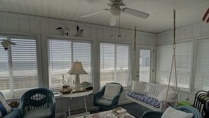 Quarto com ventilador de teto, sofá branco, duas cadeiras azuis, mesa pequena com abajur e balanço pendurado.