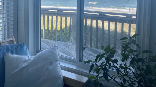 Quarto com vista para a praia através de uma grande janela, um deck de madeira e um vaso com planta.