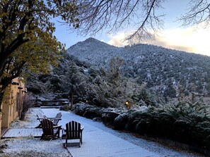 A snow-covered outdoor seating area with Adirondack chairs and a wooden table, surrounded by snow-laden trees and a mountain in the background. - Cozy Adobe Condo near Taos Ski Valley - Hacienda de Valdez (Valdez)