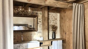 Een badkamer met een stenen muur, houten balken, een marmeren wastafel, een spiegel met decoratieve lijst, een vaas met gedroogde bloemen en een handdoek aan een rek.