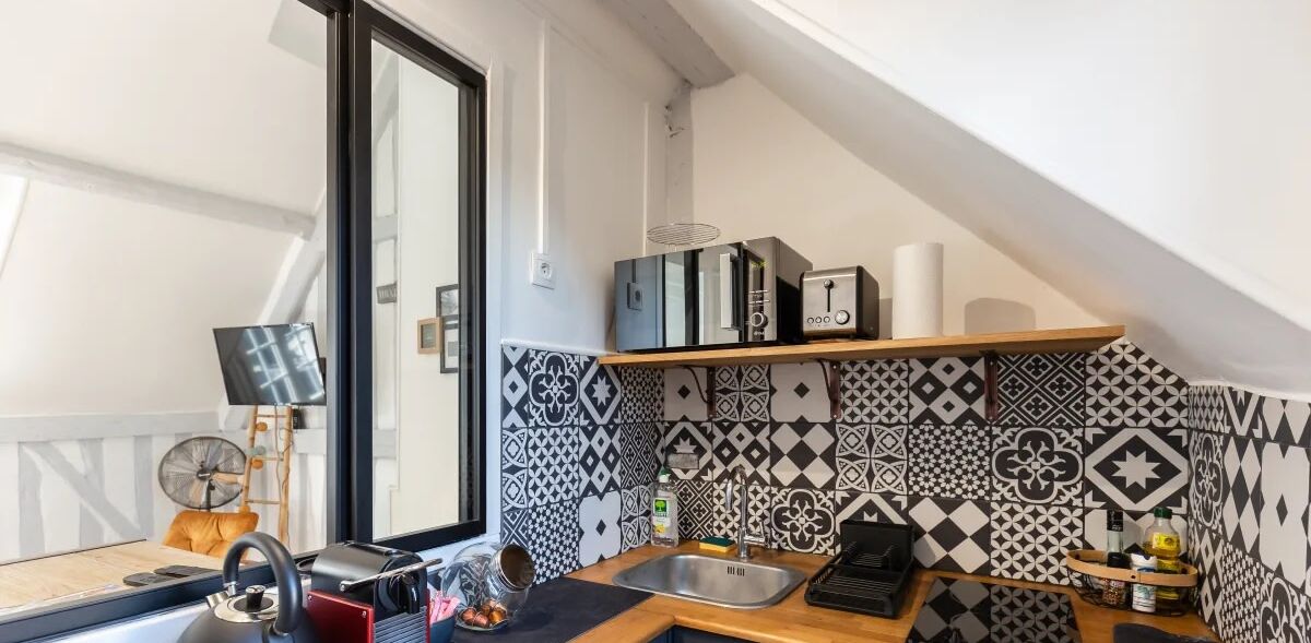 A compact kitchen with a black and white patterned backsplash, a wooden countertop, a sink, and a small shelf with kitchen appliances.