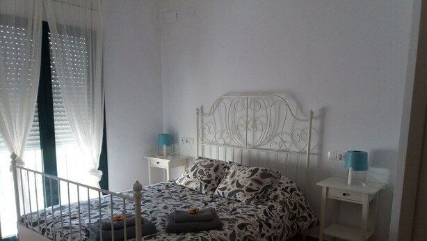 A bedroom with a bed, white nightstands, a blue lamp, and a window with curtains.