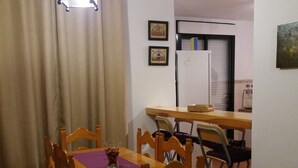 A dining area with a wooden table and chairs, a hanging light fixture, and a kitchen in the background.