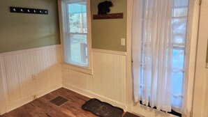 Ein Zimmer mit Holzboden, Kleiderhaken, einem Fenster mit Jalousien, einer weißen Tür mit Vorhang und einem kleinen Regal mit einem dekorativen Gegenstand.