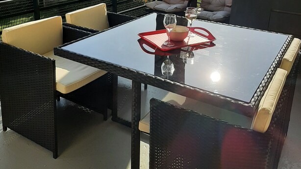 Een terras buiten met een tafel met glazen blad, zachte stoelen en een rode dienblad.