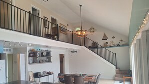 Eine geräumige Küche mit einer zentralen Kochinsel, einem Essbereich mit Stühlen, einer Treppe, die in einen oberen Bereich führt, und Blick ins Freie.