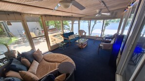 Ein geräumiger Wohnbereich mit einem großen runden Sofa, einem Couchtisch und einem Esstisch. Mehrere Stühle und ein Deckenventilator sind vorhanden. Große Fenster bieten einen Blick ins Freie.