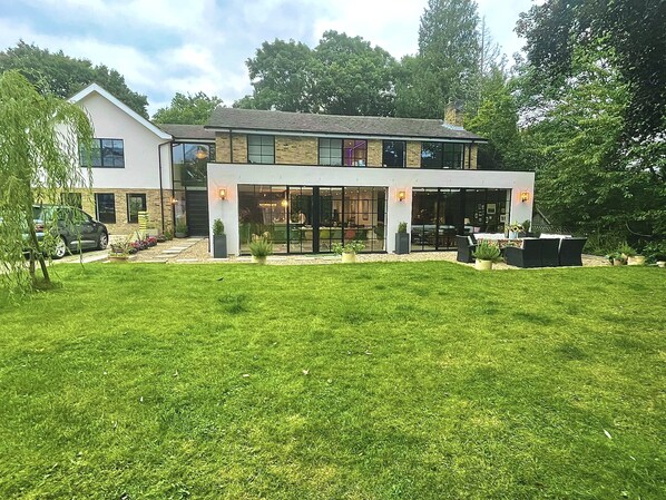 A modern two-story house with a large front yard, a patio with outdoor furniture, and a car parked in the driveway. - Grand open plan modern home with heaps of light, and close to London (Kingston upon Thames)