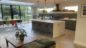 A modern kitchen with a large island, bar stools, and a dining area with a table and chairs. - Grand open plan modern home with heaps of light, and close to London (Kingston upon Thames)