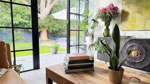 A modern living room with a large window, a wooden coffee table, a sofa, and a variety of plants and books. - Grand open plan modern home with heaps of light, and close to London (Kingston upon Thames)