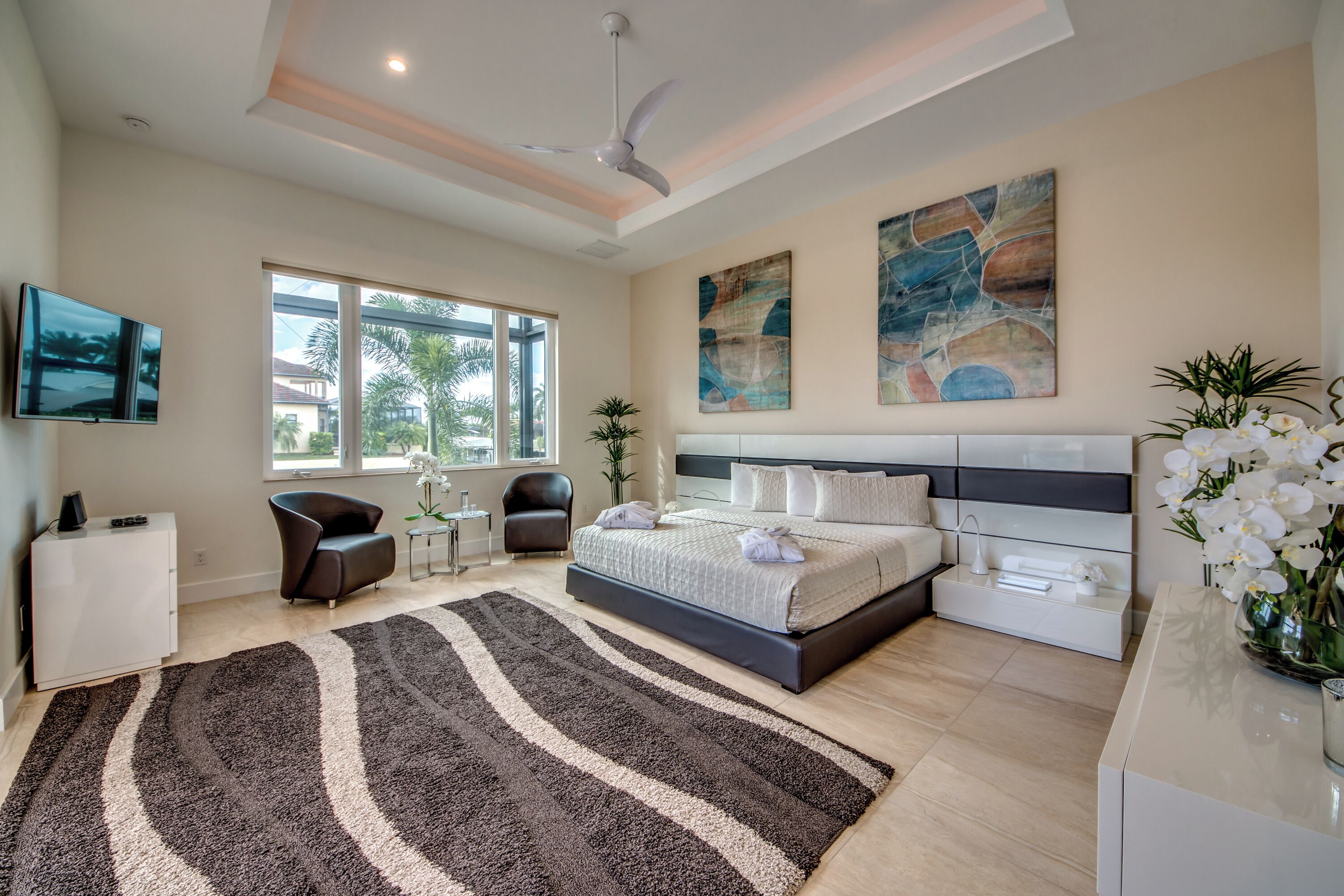 A modern bedroom with a large bed, a flat-screen TV, a ceiling fan, and artwork on the walls.
