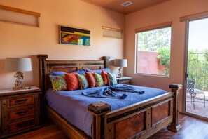 Ein Schlafzimmer mit einem hölzernen Bett, farbenfrohen Kissen, einem Bild an der Wand und einem Fenster mit Blick auf Grünflächen.