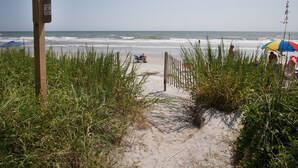 Een zandpad langs het strand dat naar de oceaan leidt, met een houten bord en een strandparasol op de achtergrond.