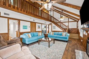Ein geräumiges Wohnzimmer mit hohen Decken, Holzbalken, einer Treppe und einem Fernseher.