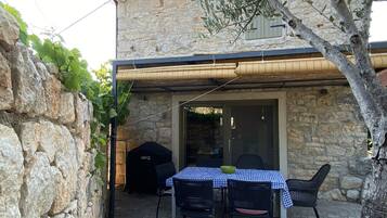 En bygning i sten med en overdækket terrasse, der har et bord, stole, grill og udsigt over det omkringliggende landskab.