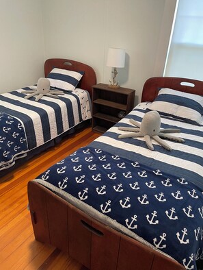 A room with two beds, each with a striped navy and white bedding featuring anchor patterns. There is a nightstand with a lamp between the beds.