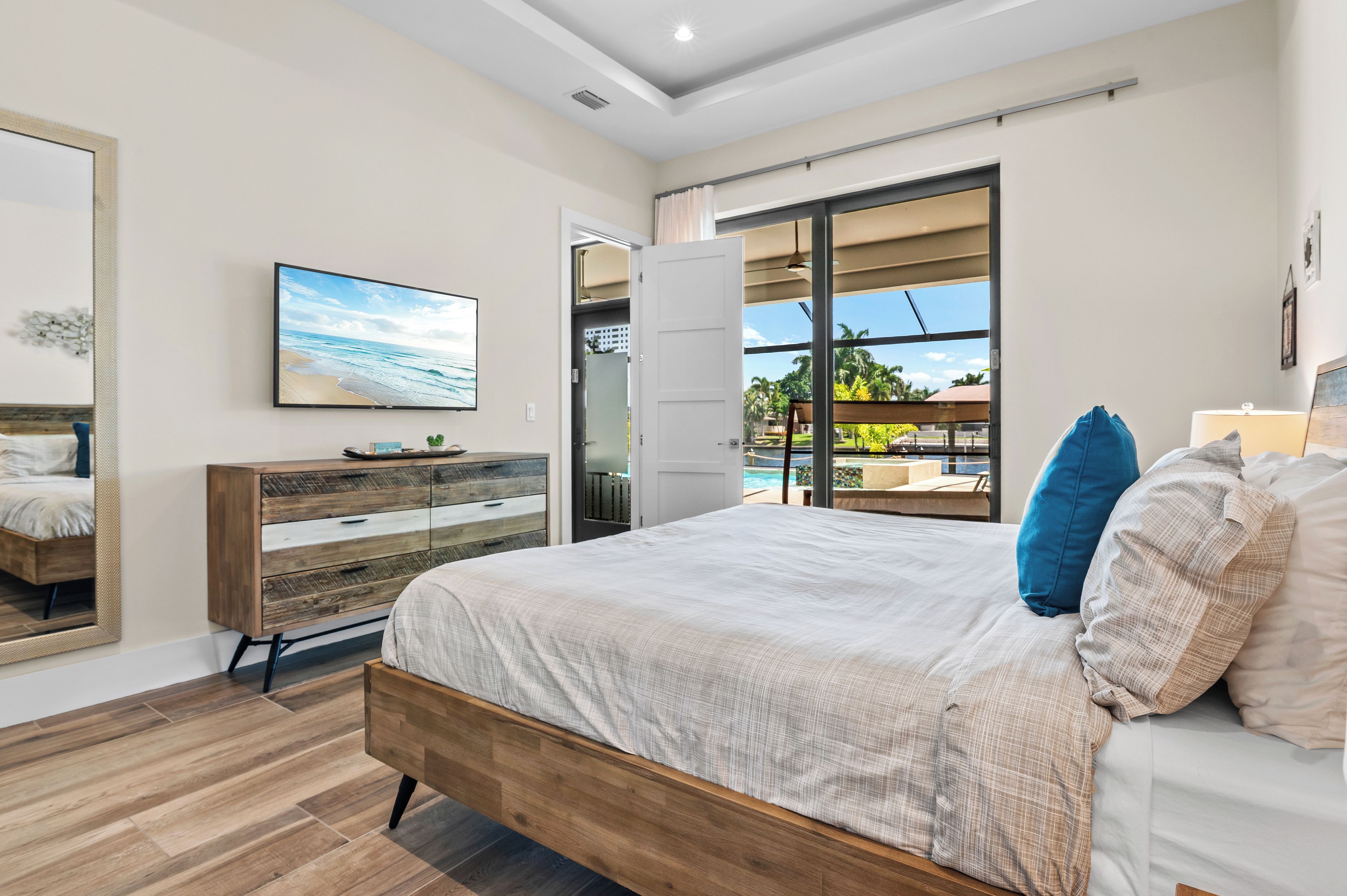 Ein Schlafzimmer mit Bett, Kommode, Fernseher und Blick auf Pool und Palmen.