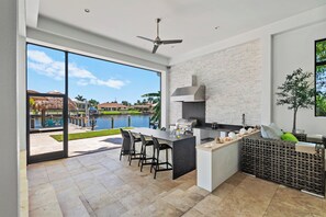 A modern kitchen with a large island, a built-in grill, and a view of a pool and palm trees.