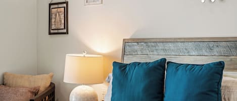 A neatly made bed with two teal pillows, a wooden headboard, a bedside lamp, and a wall-mounted decorative piece.