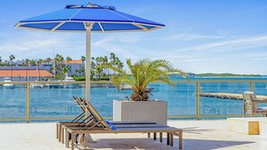 A sunlit outdoor lounge area with a blue umbrella, lounge chairs, and a palm plant.