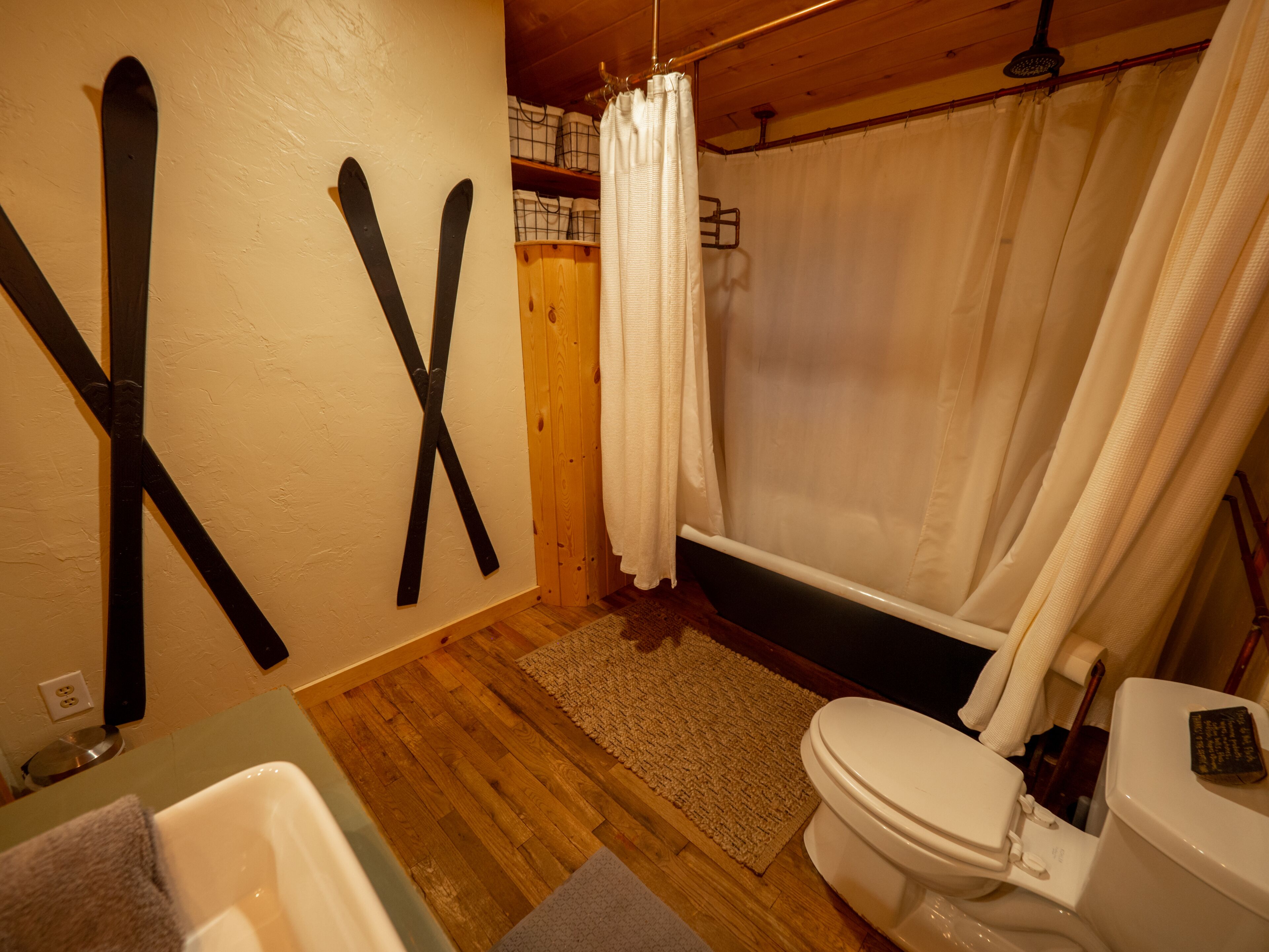 Et badeværelse med trægulv, et hvidt toilet, en bruseforhæng og to ski monteret på væggen.