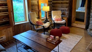 Interior acogedor de una cabaña de madera con una mesa, dos sillas, una lámpara y vistas al exterior.