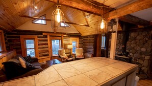 Interior de una cabaña de troncos con una isla de cocina grande y con baldosas, vigas de madera en el techo y una chimenea de piedra.