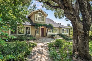 A house with a stone pathway leading to the entrance, surrounded by lush greenery and a large tree.
